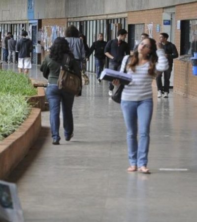 Estudantes circulam pelo corredor de uma universidade pública brasileira, carregando mochilas e materiais de estudo.