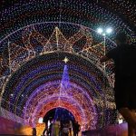Foto noturna de uma grande árvore de Natal iluminada com luzes coloridas, montada em via pública, com decoração natalina ao redor e clima festivo.