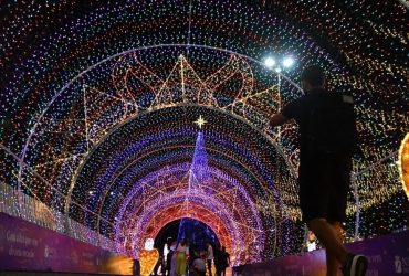 Foto noturna de uma grande árvore de Natal iluminada com luzes coloridas, montada em via pública, com decoração natalina ao redor e clima festivo.