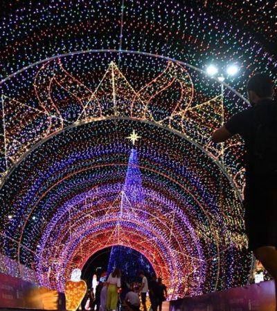 Foto noturna de uma grande árvore de Natal iluminada com luzes coloridas, montada em via pública, com decoração natalina ao redor e clima festivo.