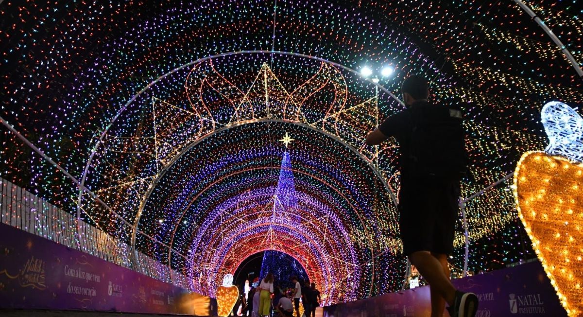 Foto noturna de uma grande árvore de Natal iluminada com luzes coloridas, montada em via pública, com decoração natalina ao redor e clima festivo.