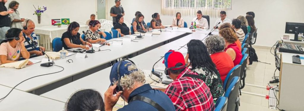 A imagem mostra uma reunião formal entre representantes da Universidade Federal Rural do Semi-Árido e do Instituto Nacional de Ciências Agrícolas – INCA, de Cuba. Os participantes estão sentados ao redor de uma mesa retangular ampla, equipada com microfones individuais, documentos e blocos de anotação. Alguns utilizam fones de ouvido, indicando a presença de tradução simultânea, o que reforça o caráter internacional do encontro. Ao fundo da sala, sobre uma mesa lateral, aparecem pequenas bandeiras do Brasil e de Cuba, simbolizando a parceria entre as instituições. O ambiente é claro, organizado e com disposição típica de reuniões institucionais, sugerindo um momento de diálogo oficial, alinhamento de ações e planejamento de cooperação acadêmica.