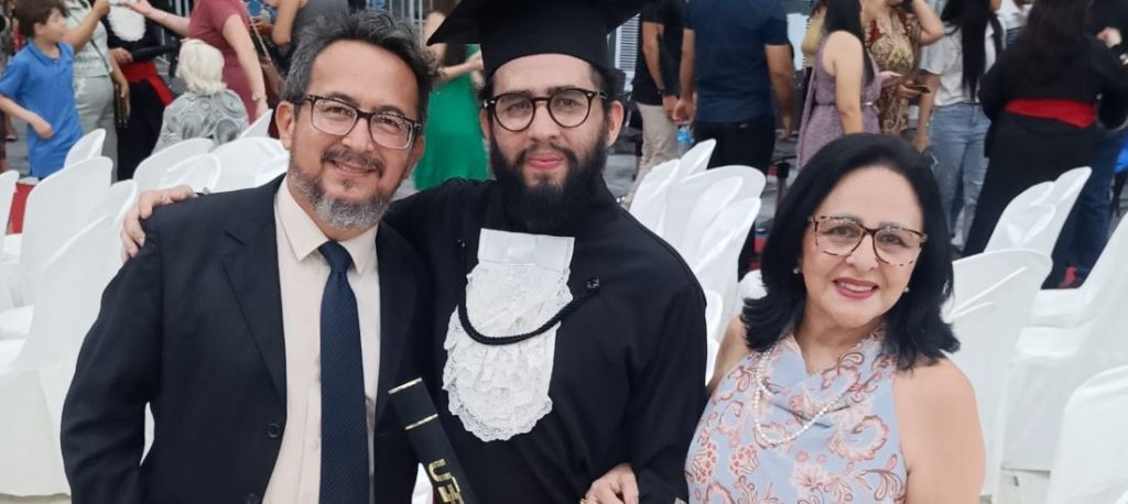 A foto registra um momento de celebração na colação de grau do filho do jornalista Passos Jr. No centro, o formando aparece com beca preta, faixa branca rendada e capelo, segurando o canudo do diploma e sorrindo com expressão de conquista. Ao seu lado, os pais o abraçam com orgulho: o pai veste terno escuro e gravata, e a mãe usa vestido claro com estampa delicada e óculos, ambos visivelmente emocionados. Ao fundo, cadeiras brancas e outras pessoas reunidas reforçam o clima festivo e solene da cerimônia, marcada por alegria, união familiar e sentimento de dever cumprido.