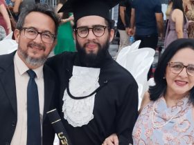A foto registra um momento de celebração na colação de grau do filho do jornalista Passos Jr. No centro, o formando aparece com beca preta, faixa branca rendada e capelo, segurando o canudo do diploma e sorrindo com expressão de conquista. Ao seu lado, os pais o abraçam com orgulho: o pai veste terno escuro e gravata, e a mãe usa vestido claro com estampa delicada e óculos, ambos visivelmente emocionados. Ao fundo, cadeiras brancas e outras pessoas reunidas reforçam o clima festivo e solene da cerimônia, marcada por alegria, união familiar e sentimento de dever cumprido.