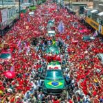 Multidão vestida de vermelho participa de uma manifestação política em apoio a Lula, com bandeiras e veículos cercados por eleitores em uma rua.