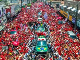 Multidão vestida de vermelho participa de uma manifestação política em apoio a Lula, com bandeiras e veículos cercados por eleitores em uma rua.