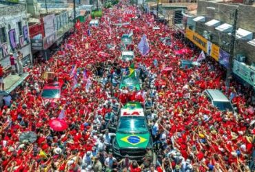 Multidão vestida de vermelho participa de uma manifestação política em apoio a Lula, com bandeiras e veículos cercados por eleitores em uma rua.