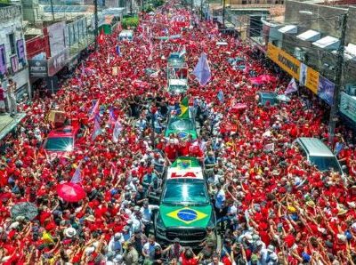 Multidão vestida de vermelho participa de uma manifestação política em apoio a Lula, com bandeiras e veículos cercados por eleitores em uma rua.