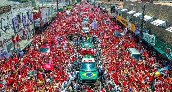 Multidão vestida de vermelho participa de uma manifestação política em apoio a Lula, com bandeiras e veículos cercados por eleitores em uma rua.