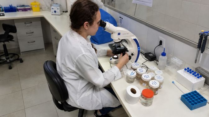 Pesquisadora observa uma amostra em um microscópio óptico em laboratório, com frascos e materiais de pesquisa sobre a bancada.