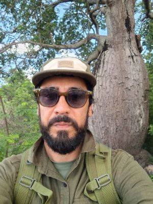 Jaqueiuton Jorge de barba e bigode pretos, usando boné bege, óculos escuros, camisa verde-oliva e mochila, posando diante de uma árvore de tronco largo em uma área arborizada.