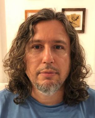 Adriano Caliman, um homem de cabelos longos, ondulados e castanhos, com barba grisalha no queixo e bigode, usando camiseta azul e olhando diretamente para a câmera em ambiente interno.