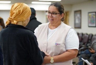 Profissional de saúde conversa com paciente usando lenço na cabeça em sala de espera.